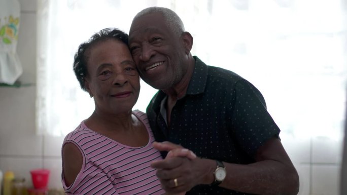 一对快乐的巴西黑人夫妇在家里的厨房里跳舞。非裔退休老人和女人脸挨着脸