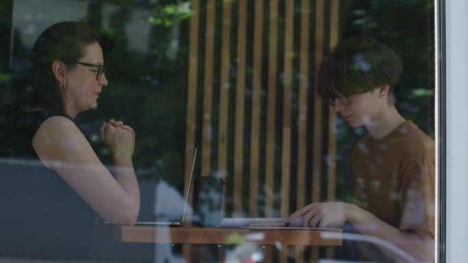 女人在咖啡馆里和年轻的男学生说话，透过窗户看窗外，手里拿着报纸