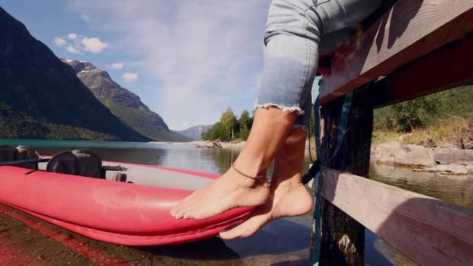 女人的脚在一个被群山环绕的原始湖泊上，她坐在木码头上，红色的独木舟在水面上