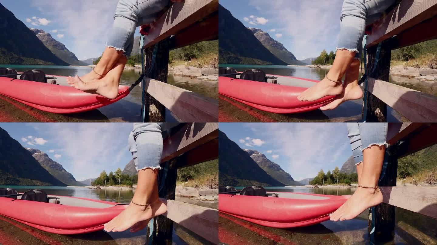 女人的脚在一个被群山环绕的原始湖泊上，她坐在木码头上，红色的独木舟在水面上