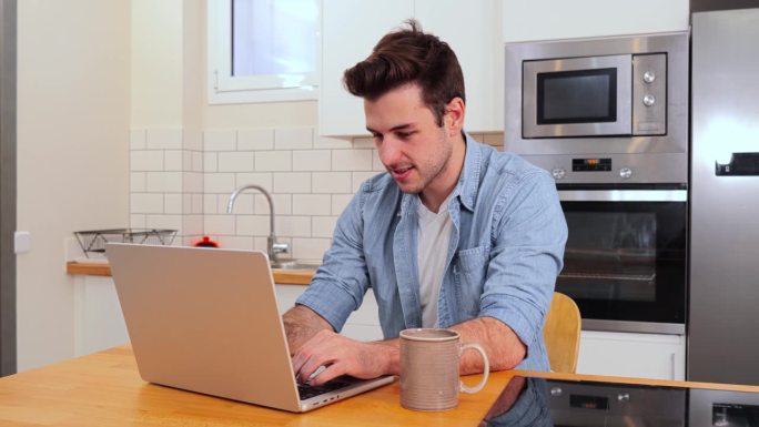 帅气的白人年轻人在家远程办公用笔记本电脑看着摄像头。快乐的男企业家正坐在厨房里用电脑微笑着使用无线网