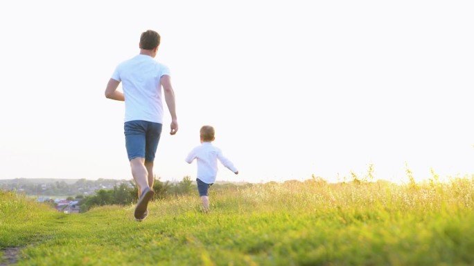 快乐的父亲和儿子在夕阳下的草地上奔跑。友好的家庭梦想生活在一起