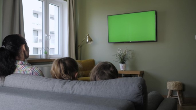 一家人坐在客厅的沙发上看电视。家庭和孩子坐在客厅沙发上的后视图，白天一起看绿色屏幕电视。