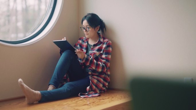 坐在客厅靠窗的年轻亚洲女子。她使用数字平板电脑并为项目创意撰写报告。