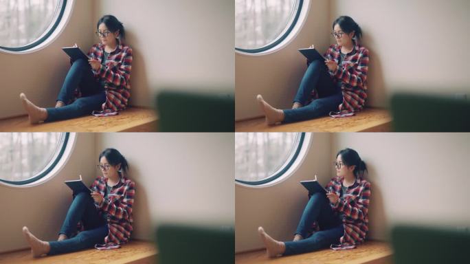坐在客厅靠窗的年轻亚洲女子。她使用数字平板电脑并为项目创意撰写报告。