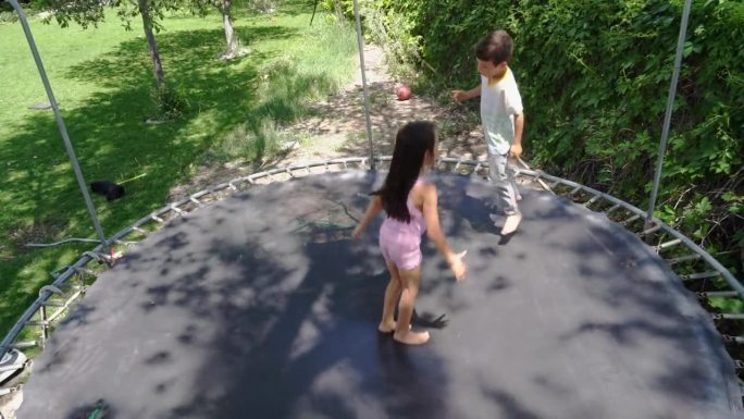 在一个大后院，一个6岁的小女孩和一个5岁的小男孩在蹦床上欢快地玩耍。绿色的草地和树木为他们提供了一个