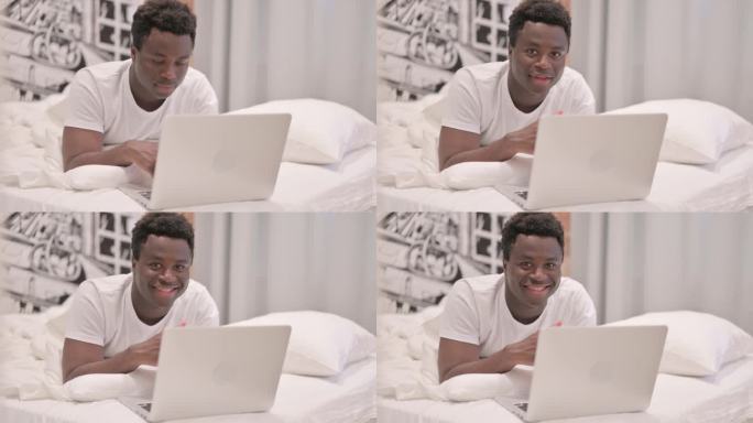 一名非洲裔美国男子趴在床上，一边用笔记本电脑工作，一边对着镜头微笑