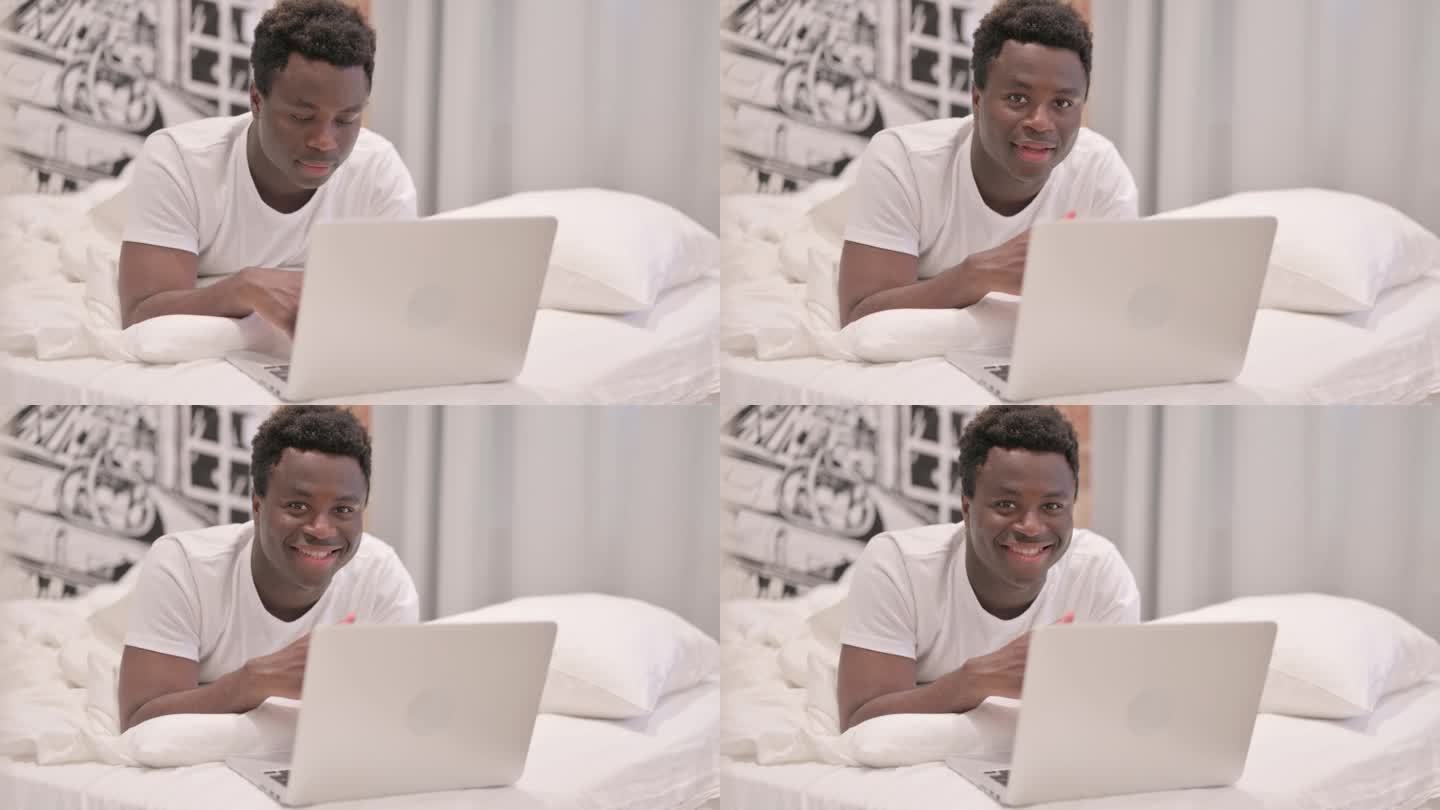 一名非洲裔美国男子趴在床上，一边用笔记本电脑工作，一边对着镜头微笑