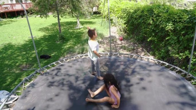 在一个绿色的院子里，蹦床活跃起来，一个6岁的小女孩和一个5岁的小男孩在蹦床上跳着，咯咯地笑着。院子里