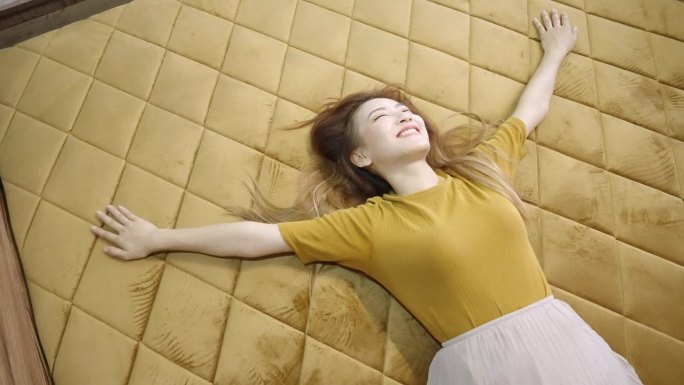 一个俏皮的亚洲女人舒服地躺在一张铺着金色天鹅绒格子羽绒被的床上，用高角度拍摄
