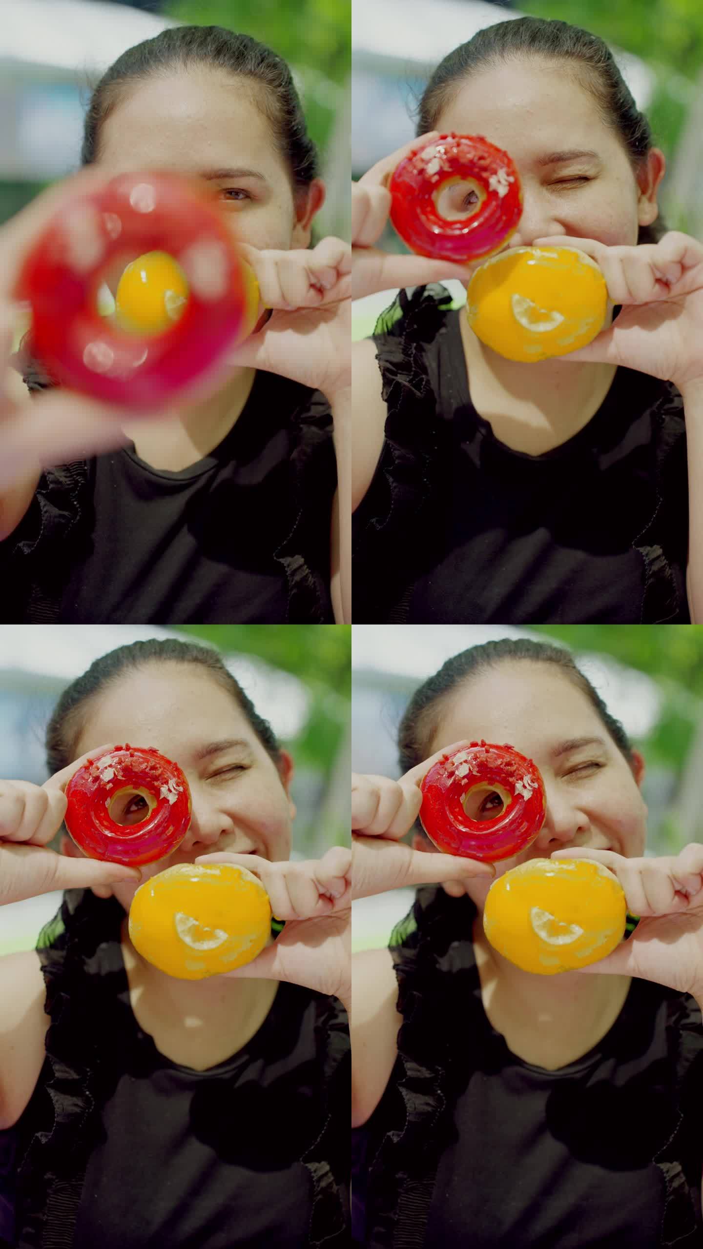 特写:看着镜头:亚洲中年妇女双手拿着两个红色和黄色的甜甜圈，和摄影师一起玩、移动甜甜圈。她的眼睛透过