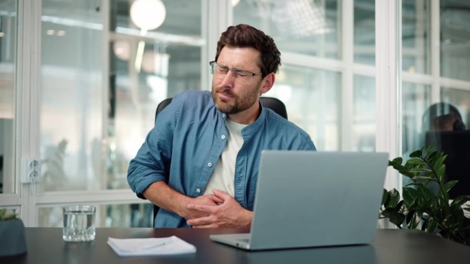 戴眼镜的年轻上班族商人男工人留胡子的男性坐在工作场所用笔记本电脑工作感到胃痛不适消化不良消化问题。胃