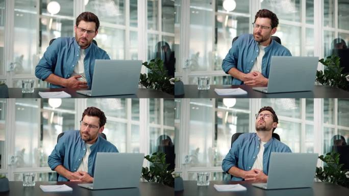 戴眼镜的年轻上班族商人男工人留胡子的男性坐在工作场所用笔记本电脑工作感到胃痛不适消化不良消化问题。胃
