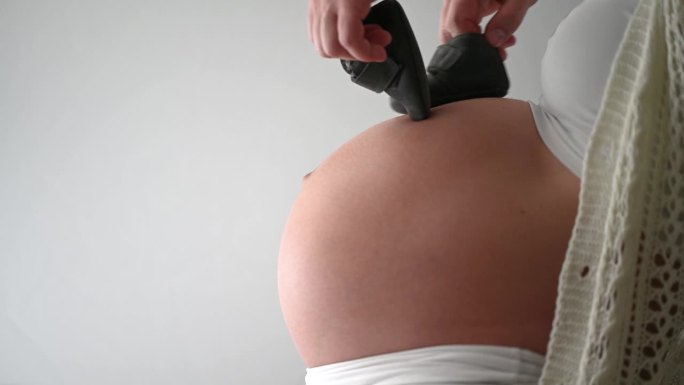 婴儿鞋在孕妇圆肚子上跳“月亮舞”的侧视图。妊娠晚期-第39周。侧视图。白色背景。自然光线。