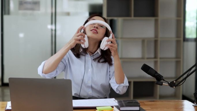一名亚洲妇女戴着耳机在笔记本电脑上录播客。女播客在她的家庭工作室制作音频播客。