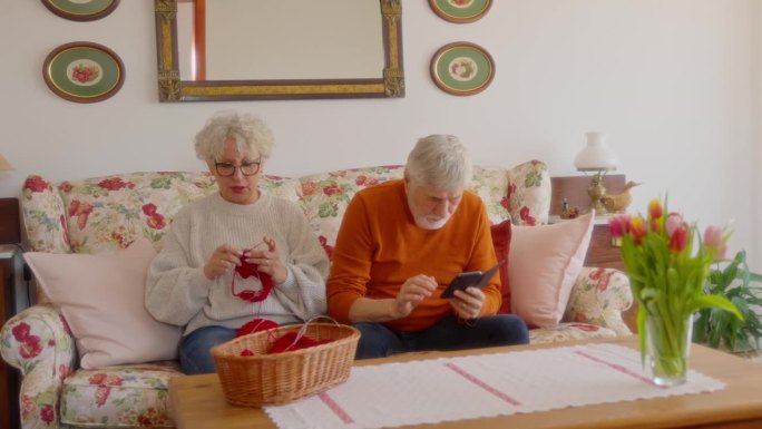 在家客厅里，老年妇女在丈夫旁边用智能手机织毛衣