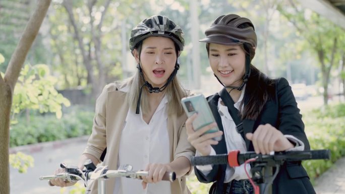 两个漂亮又活跃的亚洲女商人，一个穿着黑色西装，拿着电动滑板车和手机在城市街道上看GPS，而她的同事拿