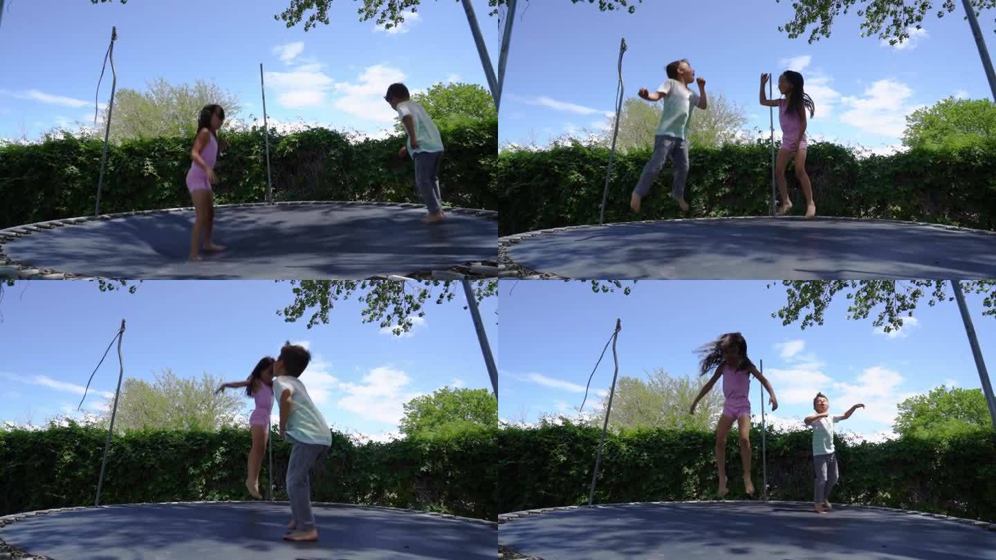 绿色院子里的蹦床为一个6岁和5岁的小女孩和男孩弹跳玩耍的愉快景象提供了背景。在夏日温暖的阳光下。