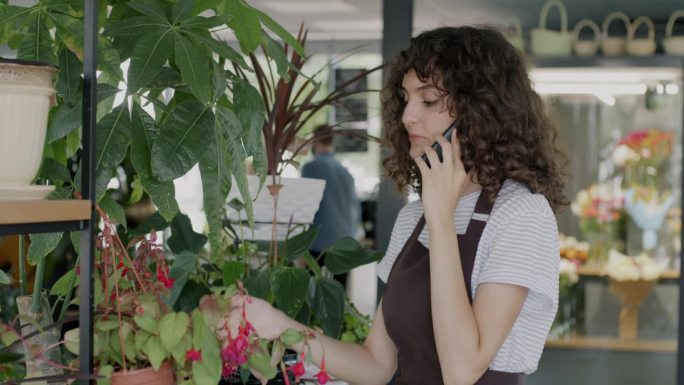 穿着围裙的花店老板年轻女子一边看着绿色植物和鲜花一边用手机和客户交谈