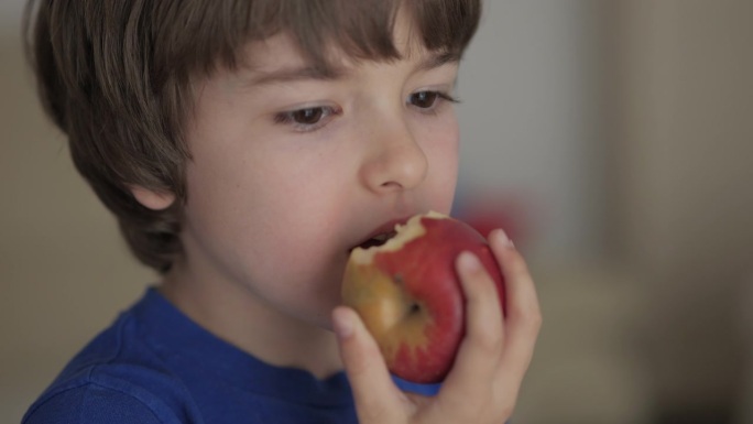 可爱的小男孩吃红苹果。孩子吃水果。小孩一边咬水果一边嚼，看着厨房里的照相机。健康饮食素食者。健康营养