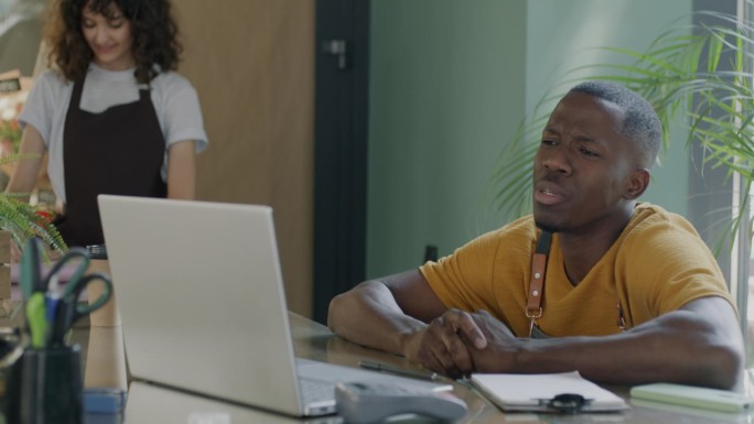 非洲裔美国男子在现代花店用笔记本电脑进行在线视频通话，女子在后台工作