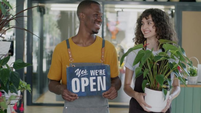 生意伙伴男女开花店举着牌子“是的，我们开门了”，绿色盆栽微笑着