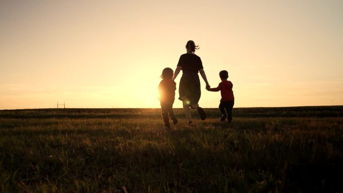 年轻家庭的剪影——妈妈和两个兄弟双胞胎男孩在露天运动场或公园跑向太阳，黄金时刻，闪耀着快乐的母亲和孩