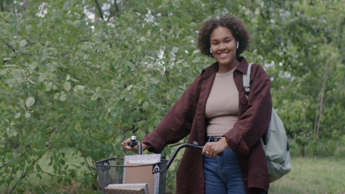 悠闲的非裔美国妇女站在绿色公园拿着自行车微笑的慢动作肖像