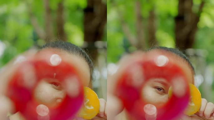 特写:看着镜头:亚洲中年妇女双手拿着两个红色和黄色的甜甜圈，和摄影师一起玩、移动甜甜圈。她的眼睛透过