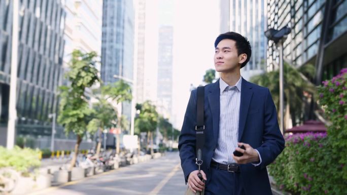 在现代城市，年轻的亚洲商人一边走在大街上一边看手机