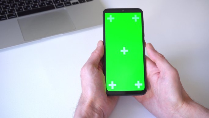 特写。男人拿着智能手机，双手卷轴，在绿色屏幕上按着他的手指。在笔记本电脑和白桌的背景上。关于人、技术