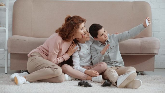 快乐微笑的家庭坐在靠近张子的地毯上，自拍，竖起大拇指，和平标志