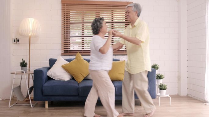 亚洲的一对老年夫妇在客厅里一边跳舞一边听音乐，甜蜜的夫妇在家里放松的时候一边享受爱情的时刻。生活方式