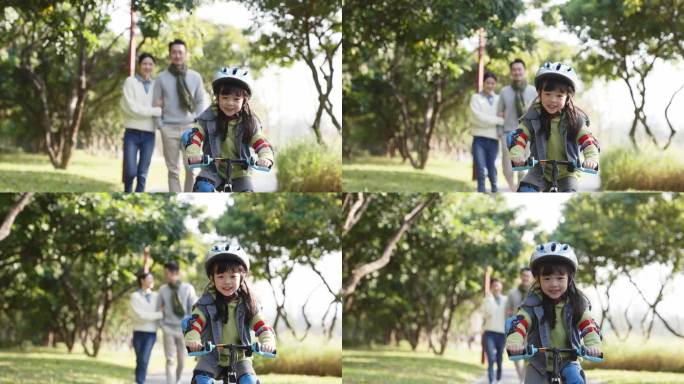亚洲小女孩骑自行车，在公园里穿着全套防护服，父母在后面看着