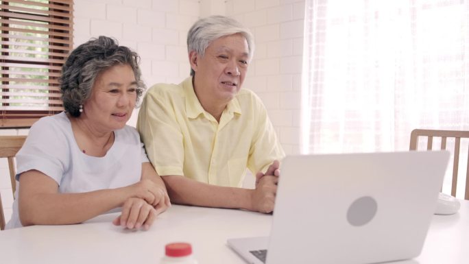 亚裔老年夫妇使用笔记本电脑与医生在客厅里谈论医学信息，夫妻俩一边躺在家里的病床上一边。家庭家庭健康概