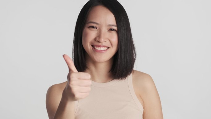 漂亮的年轻亚洲女人用食指直勾勾地对着相机，用白色的背景保持着大拇指的隔离。就像你的表情