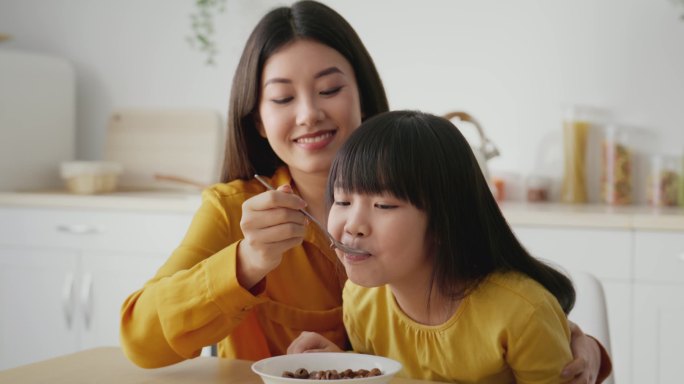 早上在家里的例行公事可爱可爱的亚裔母亲坐在厨房里，用牛奶和麦片喂女儿