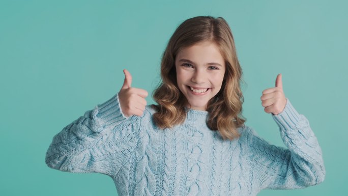 漂亮的金发少女在镜头前竖起大拇指，带着蓝色背景的微笑。身体语言概念