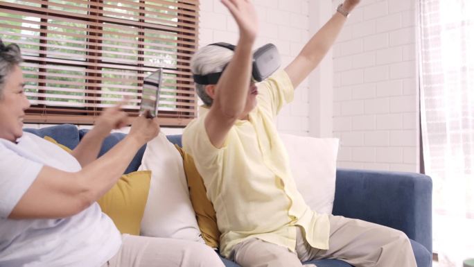 亚洲的一对老年夫妇在客厅里玩着平板电脑和虚拟现实模拟器，夫妻们在一起躺在沙发上开心地度过时光。家庭中
