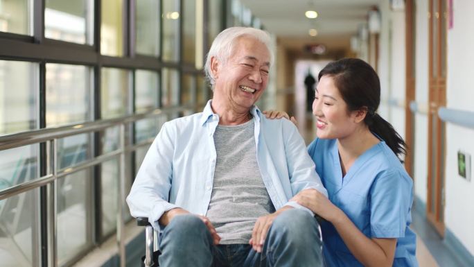 友好的亚裔工人与住在辅助生活设施走廊的坐轮椅的老人交谈