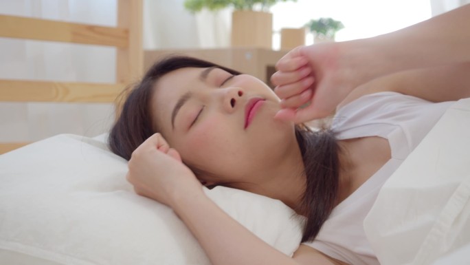 慢动作 - 美丽迷人的年轻亚洲妇女醒来，微笑和伸展她的手臂在她的床上在卧室。年轻的亚洲女性在家里使用