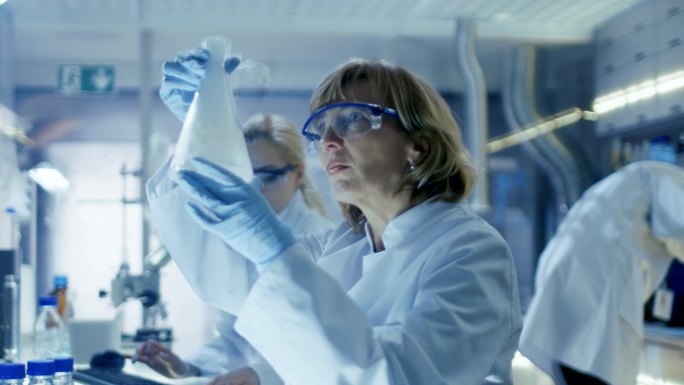 高级女化学家与液体化学品在超现代实验室工作。她坐在她的办公桌，她的同事和他们计算机上的分析数据。