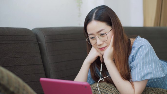 快乐的亚洲女人带着耳机，一边在平板电脑上看电影，一边在沙发上享受着放松的生活.