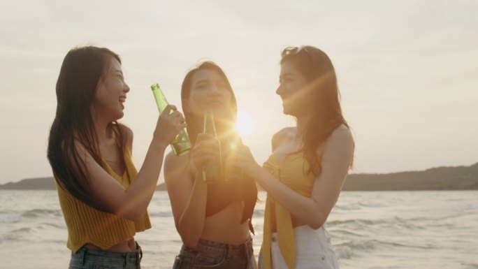 一群亚洲少女在沙滩上聚会庆祝，晚上日落时，朋友们在沙滩上愉快地喝啤酒。户外旅游假期假期夏季概念。慢动