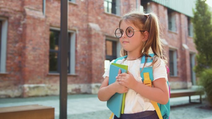戴眼镜的美丽微笑的小女生沿着小路穿过公园去上学。孩子背着书包，手里拿着课本去上学。回到学校。