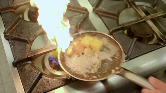 厨师在油锅中烹饪在一个商业厨房的火焰，专业厨师在一个餐馆的商业厨房在一个点燃的炉子上搅拌和翻转锅中的