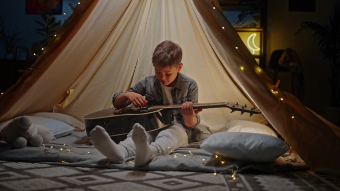 可爱的小男孩弹吉他，坐在客厅搭起的帐篷里，玩野营
