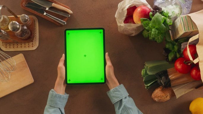 俯视图的匿名女性使用数字平板电脑与模拟绿色屏幕Chromakey显示在厨房的桌子上。女人在准备晚餐时