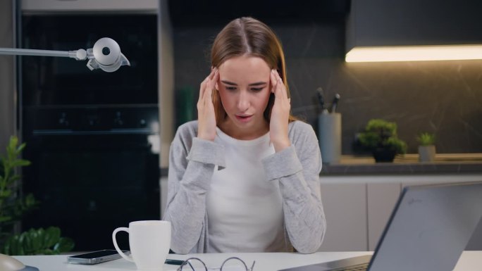 疲惫不堪的年轻女商人坐在办公室的桌子旁按摩太阳穴。女雇员学生在长时间使用笔记本电脑后感到疲劳。