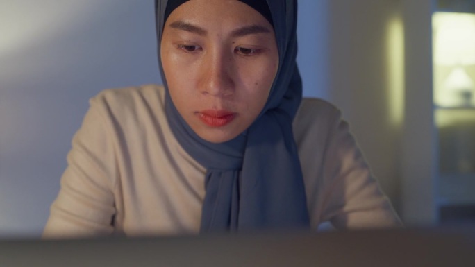 特写年轻的亚洲穆斯林自由职业者妇女戴头巾休闲布手指打字键盘笔记本电脑在家里办公室加班在晚上。小企业主